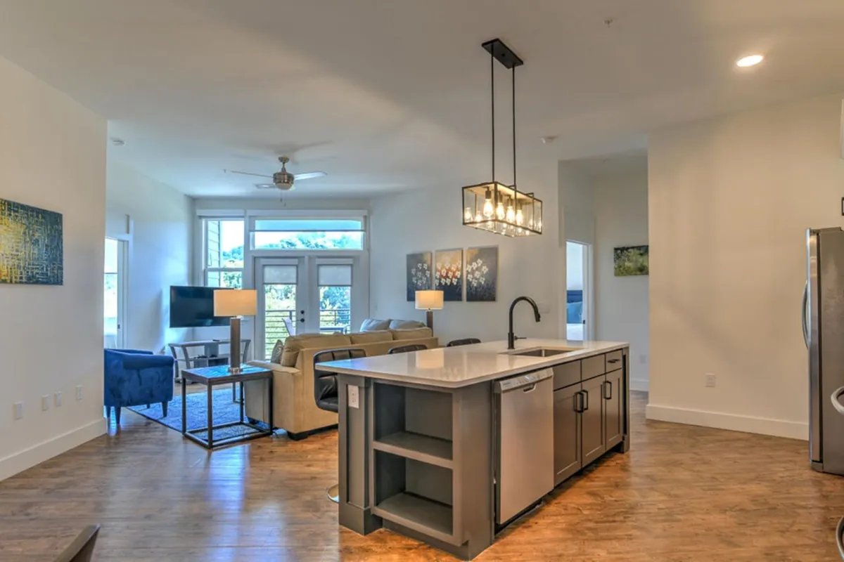 This image showcases a bright, open-plan interior space where the kitchen island seamlessly blends into the living area. The island countertop, comfortable seating, and large windows create a welcoming atmosphere filled with natural morning light, while the visible new furnishings hint at modern luxury. The space is set up for both cooking and relaxing, perfectly fitting the 'living & culinary lounge' concept.