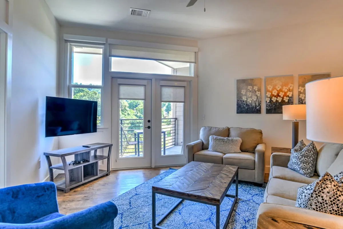 The image shows a stylish, sunlit living room with a plush sofa and coffee table, a large TV for cozy evenings, and wide balcony doors that flood the space with natural light. Contemporary artwork and fresh furnishings create a warm, welcoming atmosphere perfect for relaxing or entertaining.