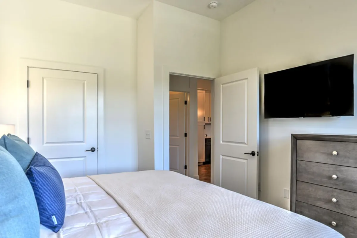 A modern bedroom featuring a plush bed dressed in blue pillows, a wall-mounted television, and a tidy dresser. The space exudes a cozy, inviting atmosphere ideal for unwinding after a day exploring Asheville.