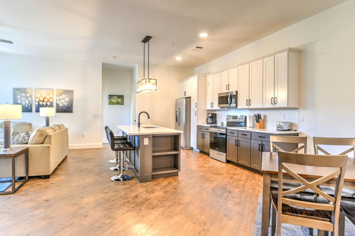 A welcoming open area combining the kitchen island, bar stools, dining table, and cozy sofa, all tied together by tasteful artwork on the wall. This perspective captures the true open-concept layout, showing how different zones flow together for dining, relaxing, or socializing. The mood is inviting and lively, perfect for family gatherings or intimate dinners.