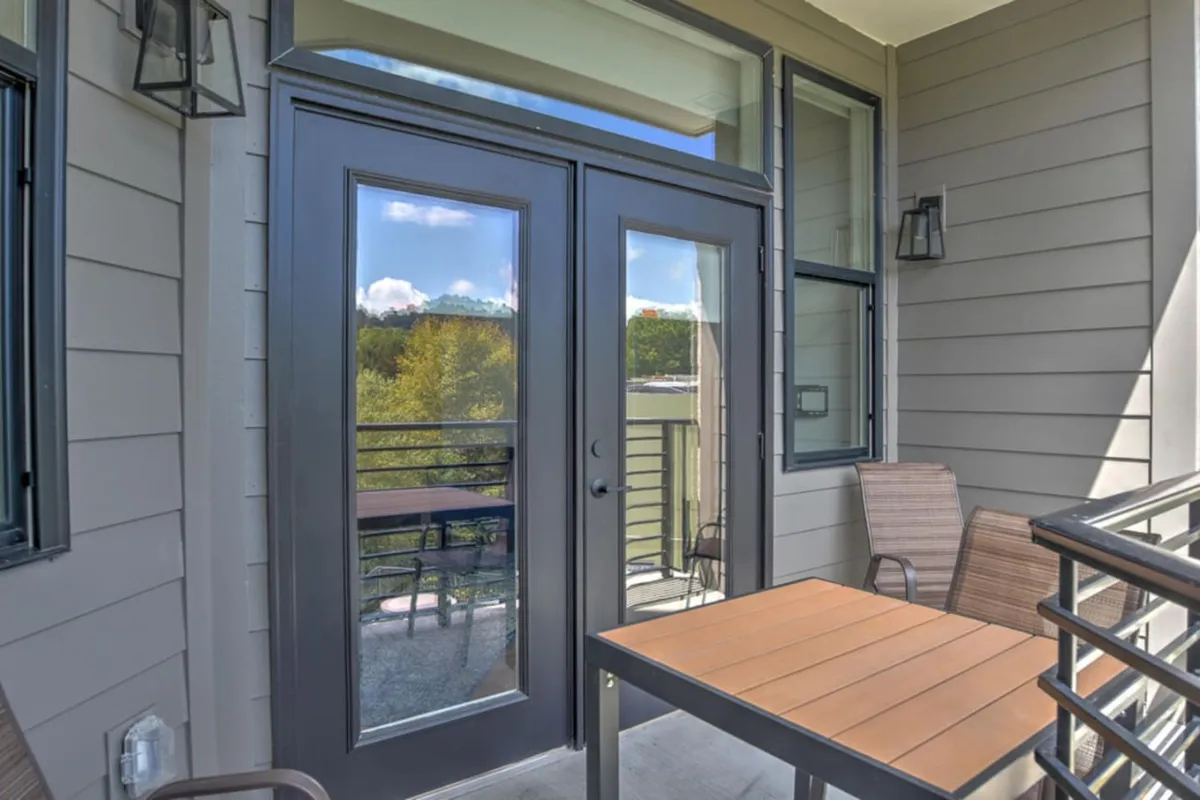 This exterior shot features double doors opening to an outdoor seating area with a table. The surrounding space is tranquil and inviting, hinting at the ease of transition from outside to inside and offering a glimpse of the lifestyle waiting within.