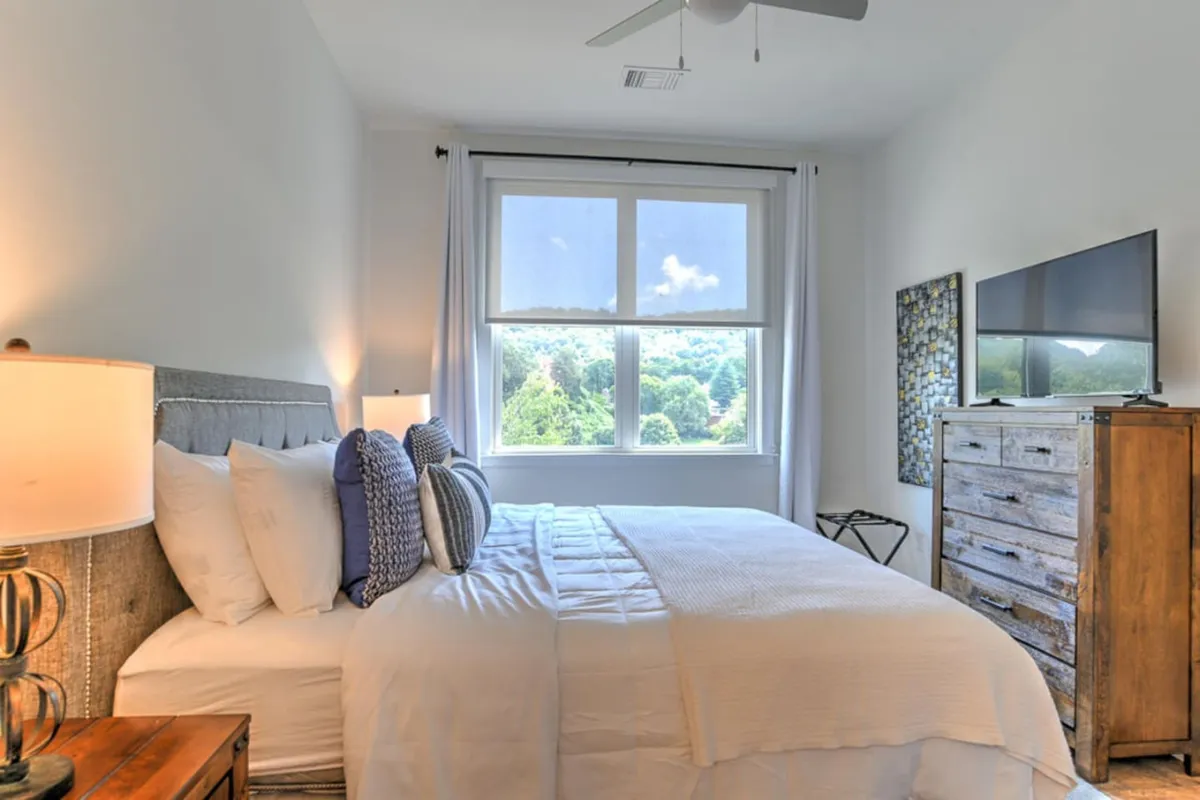 A serene bedroom with a bed positioned to enjoy the natural light streaming in from a large window, offering a glimpse of scenic mountain and city views. The setting includes a dresser and a television, enhancing comfort and relaxation.