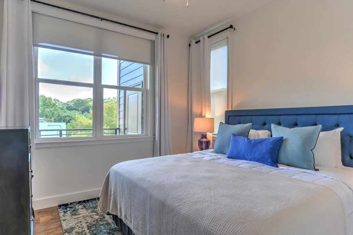A cozy bedroom with a bed accented by blue pillows, a large window offering a view, and a nightstand with a lamp. The space feels inviting and is well-lit by natural light, perfect for unwinding after a long day.