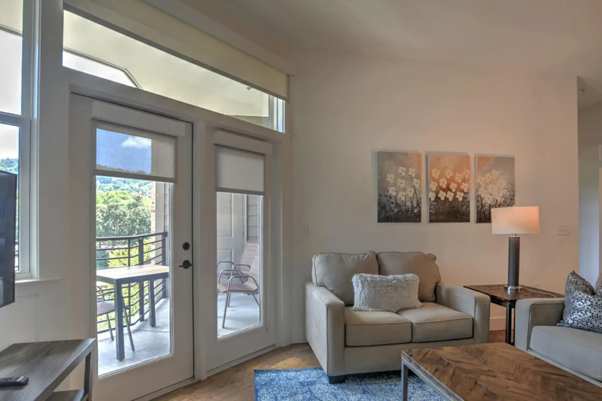 A cozy interior living space that opens directly onto the balcony through large glass doors. The room features a comfortable sofa, tasteful artwork, and ambient lighting, with the balcony visible just beyond. This indoor/outdoor connection showcases how the living space seamlessly extends to the balcony, echoing the invitation to savor both city energy and mountain tranquility from the comfort of the interior.